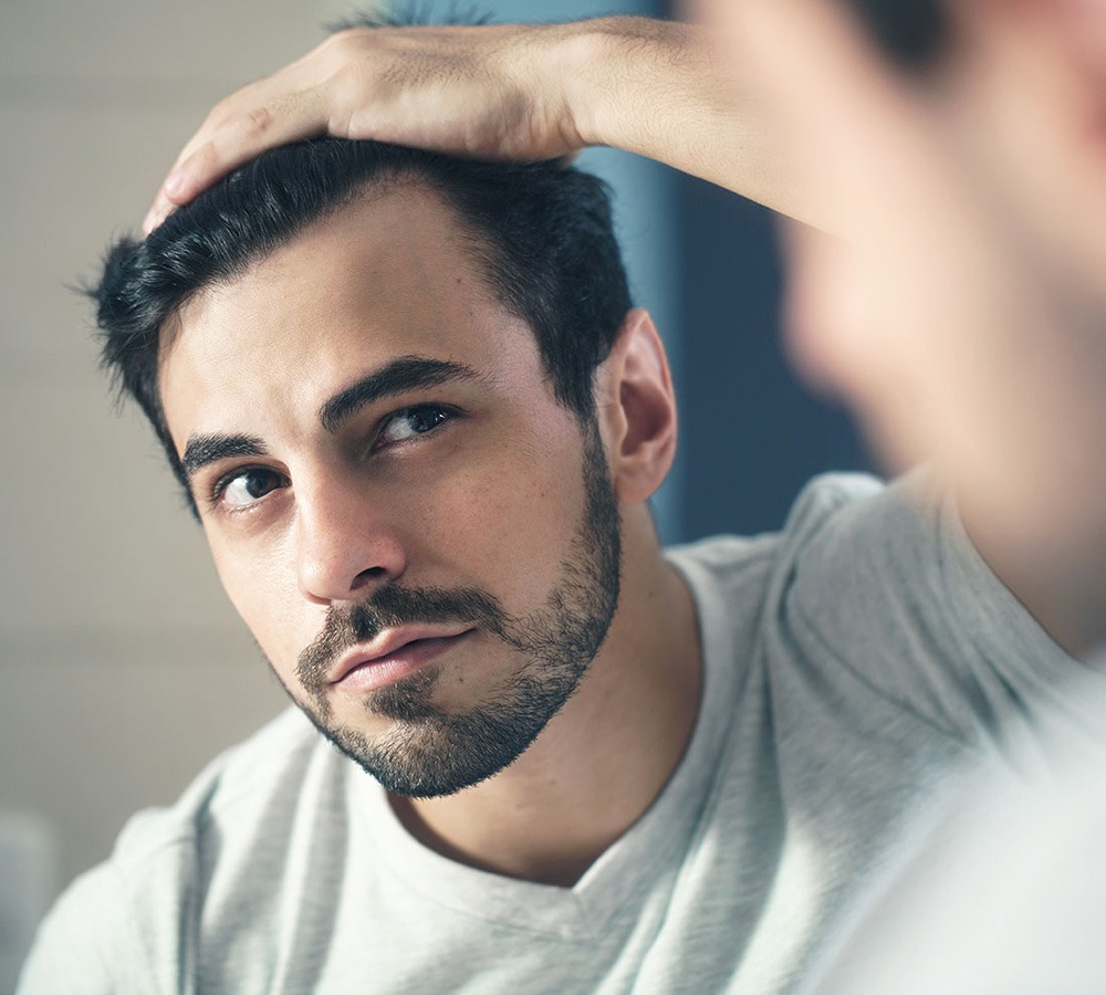 A young man with a beard examines his hair in the mirror, focusing on grooming while appearing contemplative. This image emphasizes personal care and self-reflection.
