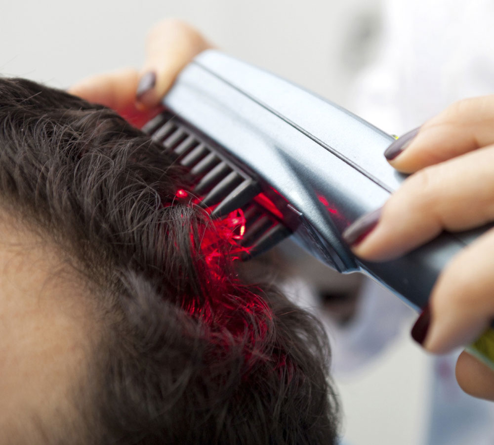 A person uses a laser comb on a client’s scalp, promoting hair growth with red light therapy. The setting appears clinical and professional.