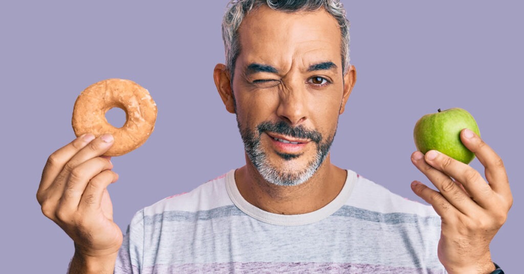 A man winks while holding a donut in one hand and a green apple in the other, highlighting the contrast between healthy and indulgent choices.