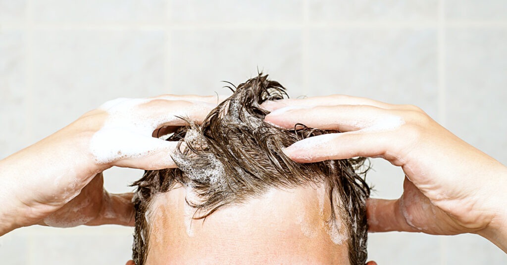 A person lathers shampoo into their hair in a shower, illustrating a hair care routine and emphasizing cleanliness.
