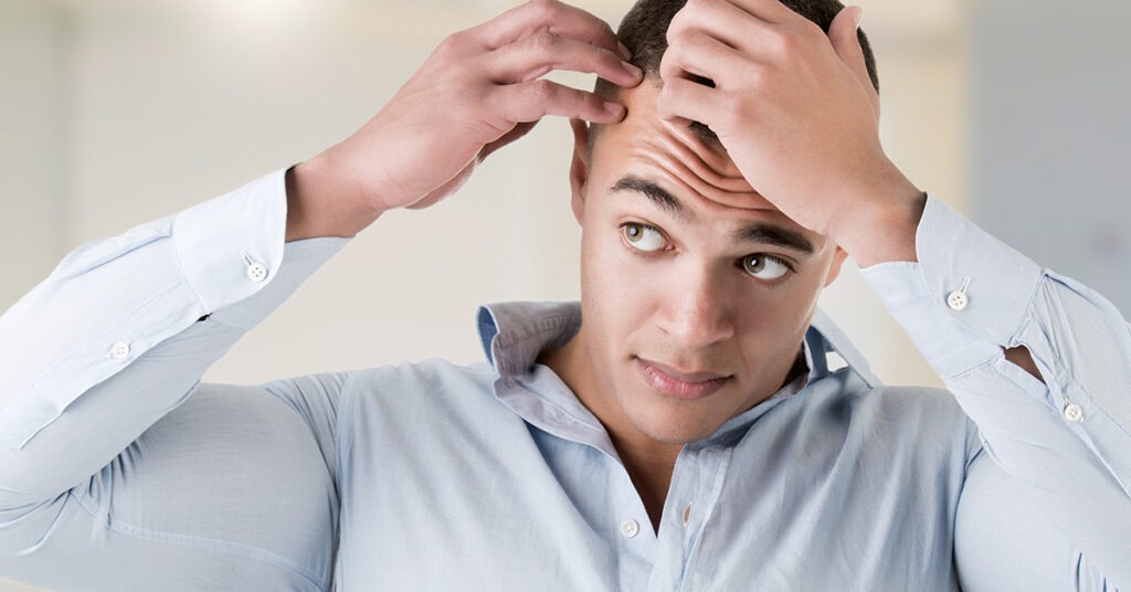 A man in a light blue shirt inspects his hairline, reflecting concerns about hair loss. The image emphasizes grooming and self-image topics.