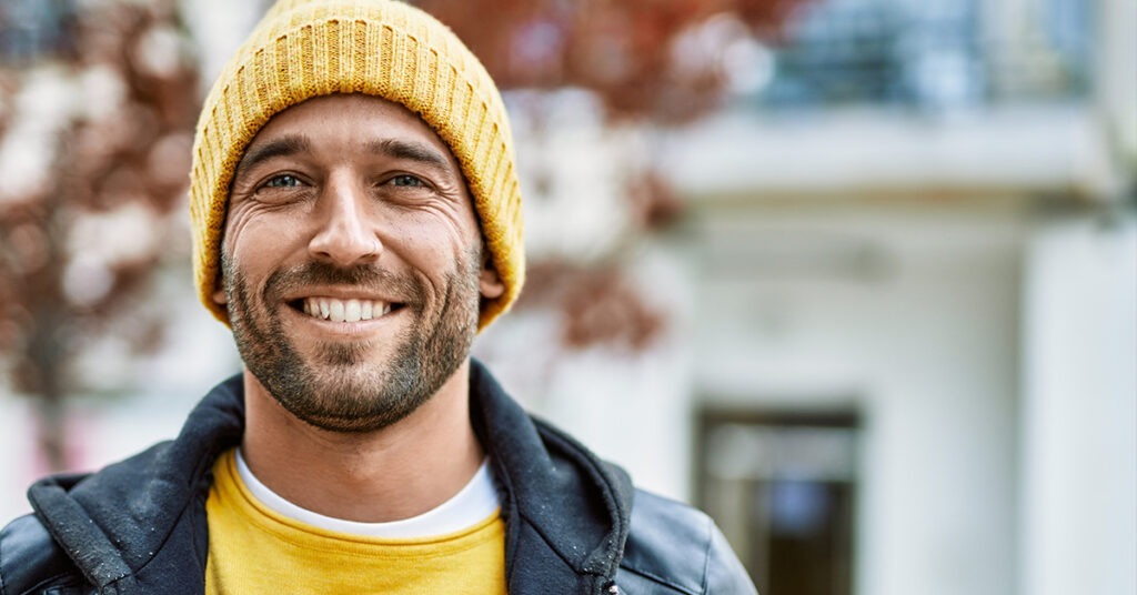Smiling man in a yellow beanie and black jacket stands outdoors, radiating warmth and friendliness in a casual urban setting.
