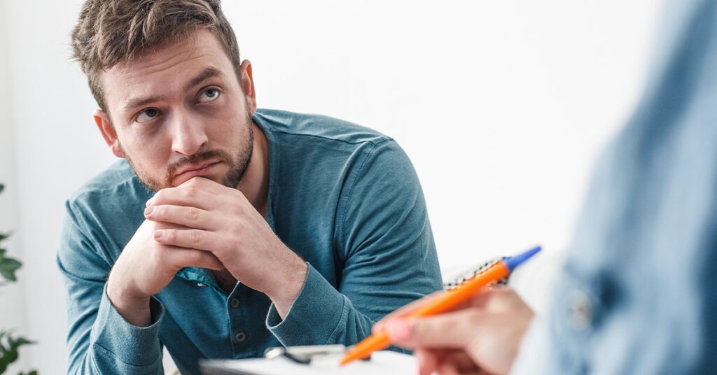A focused man with a thoughtful expression sits with clasped hands, while an out-of-focus person gestures with a pen. The scene suggests a serious conversation or counseling session.