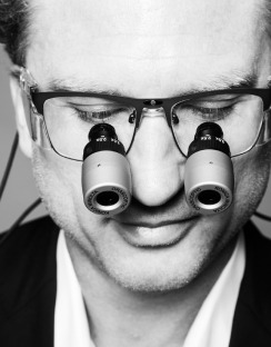 A close-up portrait of a man wearing magnifying glasses, focusing intently. His expression suggests concentration, highlighting precision and detail in his work.