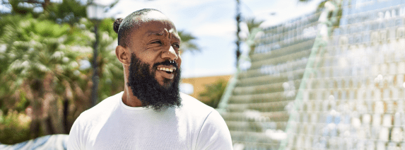 Smiling man with a beard in a white shirt stands outdoors, surrounded by palm trees and modern reflective structures, conveying a joyful and casual atmosphere.